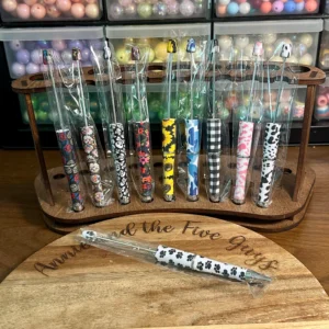 A wooden stand holds a set of Printed Beadable pens with patterned handles, in front of shelves containing various beads. A single pen lies on the wooden surface in front.