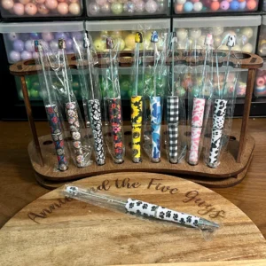 A wooden stand holds a set of Printed Beadable pens with patterned handles, in front of shelves containing various beads. A single pen lies on the wooden surface in front.