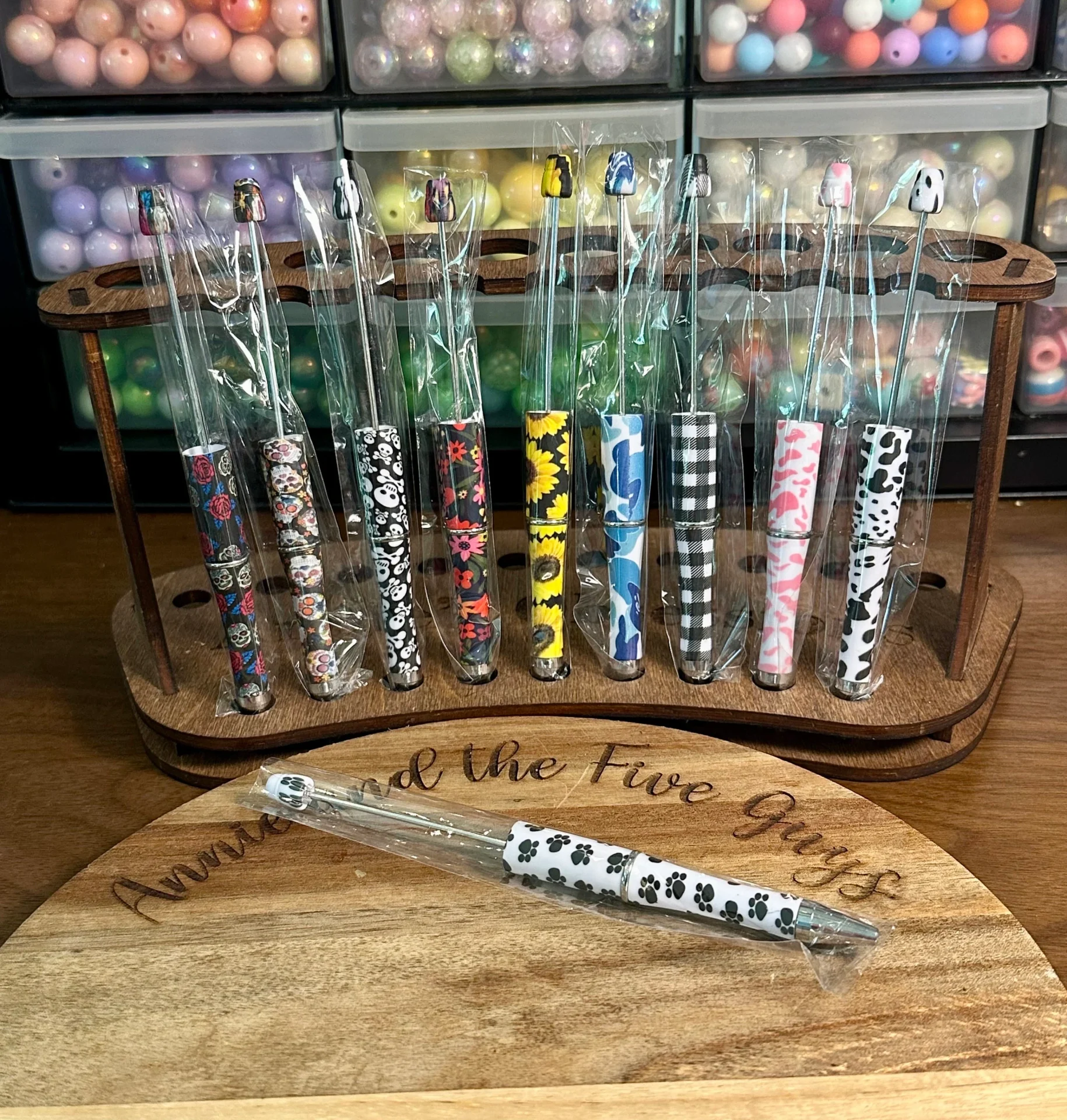 A wooden stand holds a set of Printed Beadable pens with patterned handles, in front of shelves containing various beads. A single pen lies on the wooden surface in front.