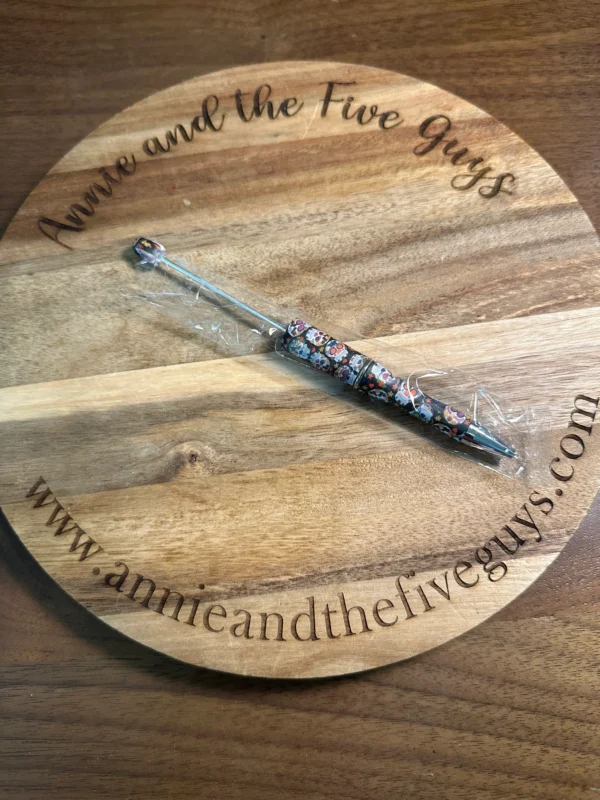 A Printed Beadable pen with intricate designs is placed on a round wooden board engraved with "Annie and the Five Guys" and a website link.