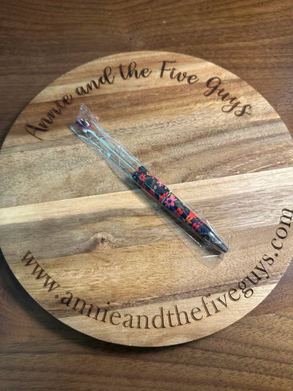 A wooden circular board with the text "Annie and the Five Guys" and a website address. In the center, a wrapped Printed Beadable pen with a colorful floral design.