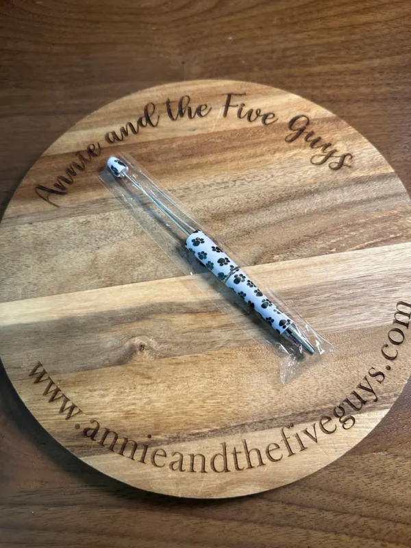 A Printed Beadable pen with a paw print design rests on a circular wooden board engraved with "Annie and the Five Guys" and the website "www.annieandthefiveguys.com".