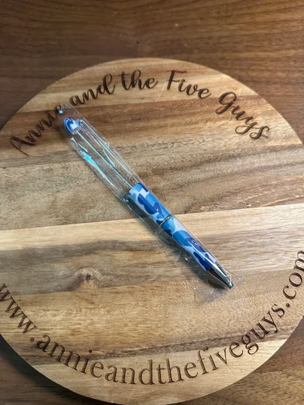 A Printed Beadable pen with blue ink lies on a wooden surface with the engraved text "Annie and the Five Guys" and the website URL www.annieandthefiveguys.com.