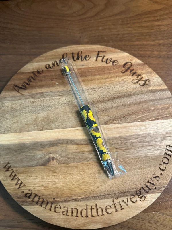 A Printed Beadable pen with a sunflower pattern is placed on a wooden surface engraved with the text "Annie and the Five Guys" and the website "www.annieandthefiveguys.com.