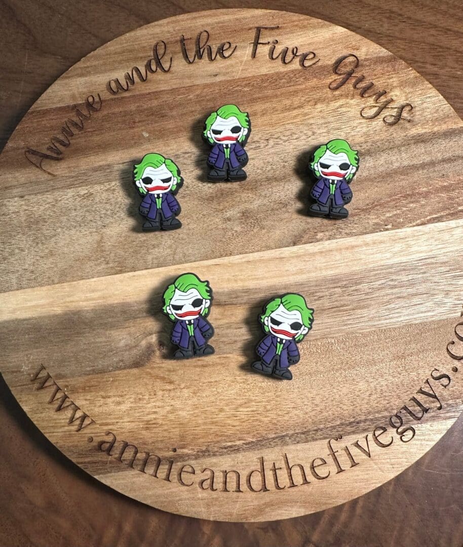 Five Jokster silicone focal beads featuring a cartoon figure with green hair and a purple suit are displayed on a round wooden board. The board has text that reads "Annie and the Five Guys" and a website URL.