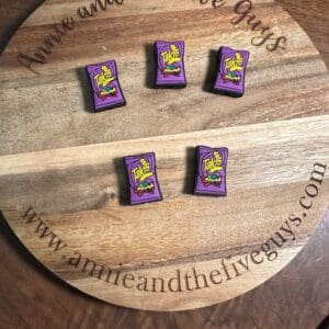 A wooden tray engraved with "Annie and the Five Guys" and a web address holds five small purple packets resembling Takis chips, arranged in two rows on a table.