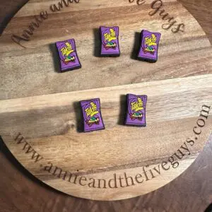 A wooden tray engraved with "Annie and the Five Guys" and a web address holds five small purple packets resembling Takis chips, arranged in two rows on a table.