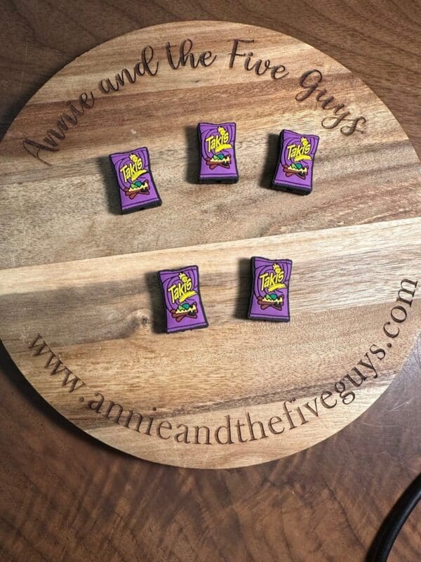 A wooden tray engraved with "Annie and the Five Guys" and a web address holds five small purple packets resembling Takis chips, arranged in two rows on a table.