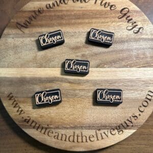 Round wooden board displaying five pins with the word "Chosen 1 Peter 2:8 silicone focal bead." The board is engraved with "Annie and the Five Guys" and a website URL.