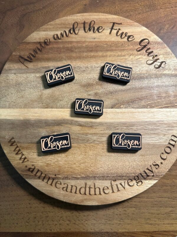 Round wooden board displaying five pins with the word "Chosen 1 Peter 2:8 silicone focal bead." The board is engraved with "Annie and the Five Guys" and a website URL.