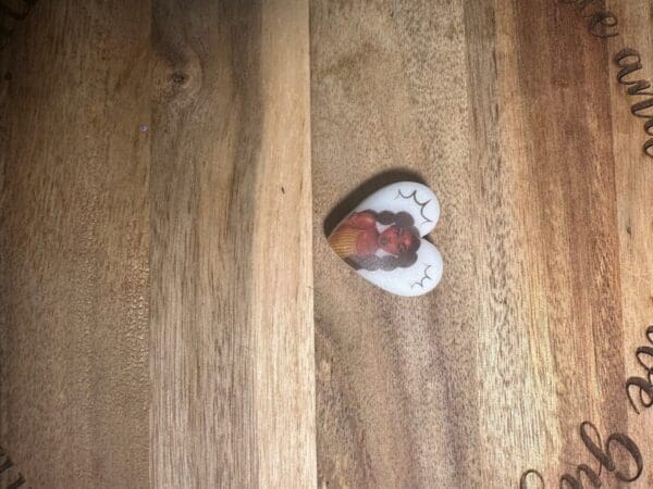 A heart-shaped pendant featuring an illustration of a person, placed on a wooden surface with faint engraved text, has been replaced with African American, African queens silicone focal beads.