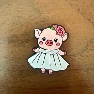 A small pin depicts a cartoon piglet wearing a white dress and a pink flower on its head, placed on a wooden surface.