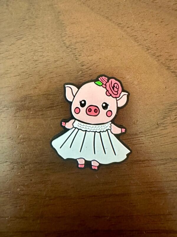 A small pin depicts a cartoon piglet wearing a white dress and a pink flower on its head, placed on a wooden surface.