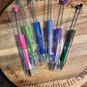 A round wooden board displaying seven Opal plastic beadable pens still in their plastic packaging. The board has the text "www.annieandthefiveguys.com" engraved on it.