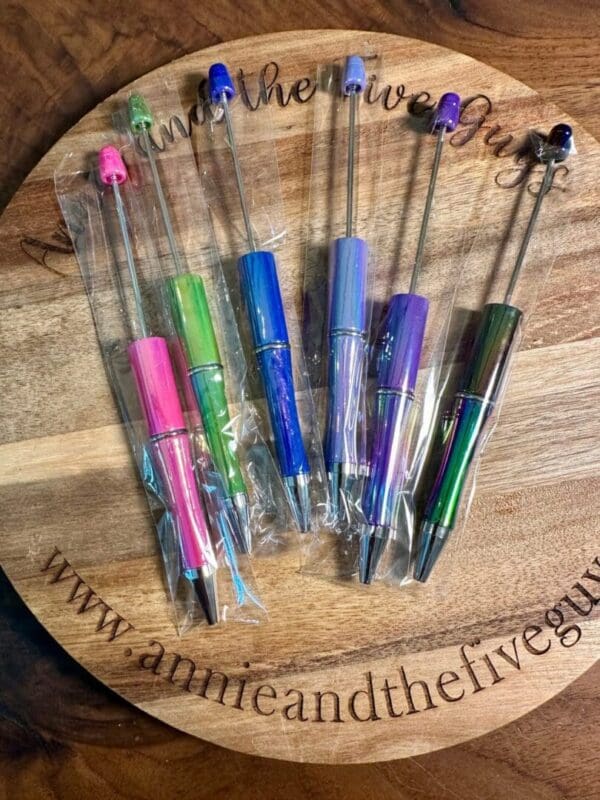 A round wooden board displaying seven Opal plastic beadable pens still in their plastic packaging. The board has the text "www.annieandthefiveguys.com" engraved on it.