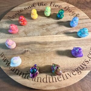 A circular wooden board with "Annie and the Five Guys" engraved around the edge. Twelve colorful glossy duckling acrylic beads are placed around the board's perimeter.