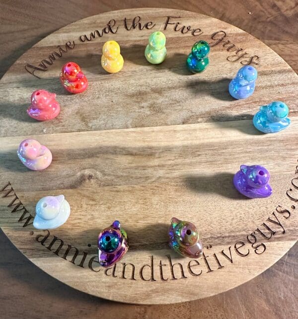 A circular wooden board with "Annie and the Five Guys" engraved around the edge. Twelve colorful glossy duckling acrylic beads are placed around the board's perimeter.