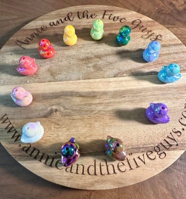 A circular wooden board with "Annie and the Five Guys" engraved around the edge. Twelve colorful glossy duckling acrylic beads are placed around the board's perimeter.