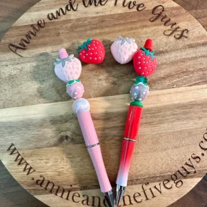 Two 3D strawberry silicone focal beads with decorative strawberry designs on top are placed on a wooden surface with the text "Annie and the Five Guys" and a website URL engraved.