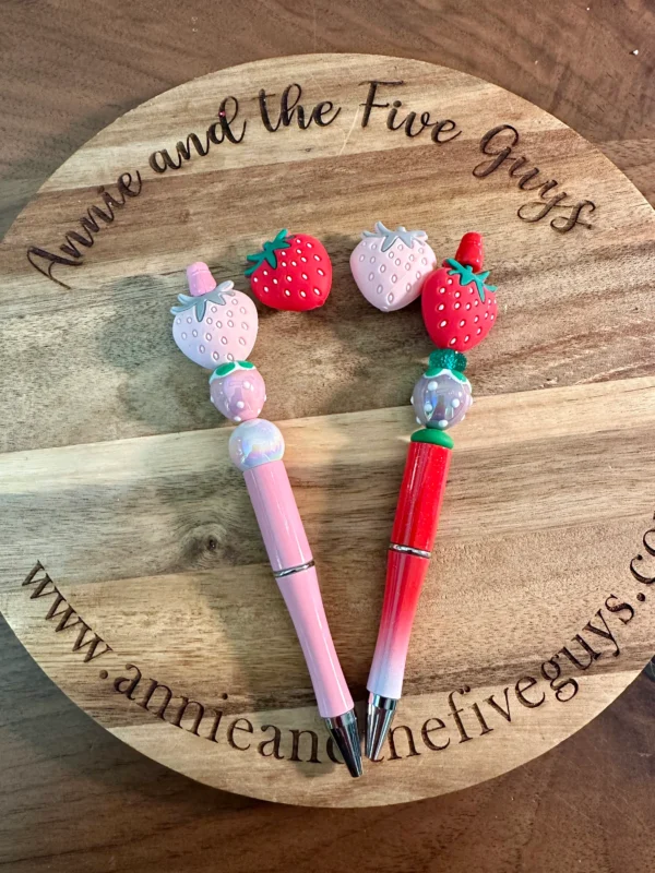 Two 3D strawberry silicone focal beads with decorative strawberry designs on top are placed on a wooden surface with the text "Annie and the Five Guys" and a website URL engraved.