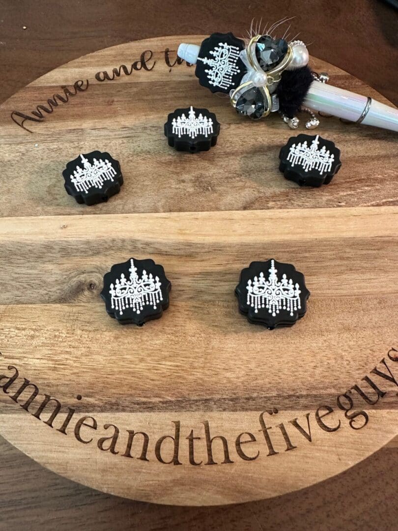 Wooden board with the words "Annie and the Five Guys" engraved, displaying black and white decorative items featuring chandelier designs. A silver writing instrument is also on the board.