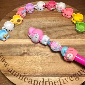 A wooden board displays small Sanrio cups resin bead arranged in an arc. A pink wand with a character on it rests in the center of the board, which is engraved with "www.annieandthefiveguys.com".