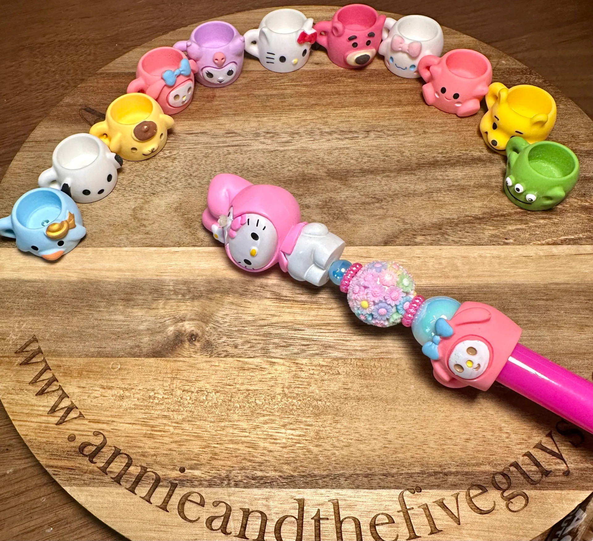 A wooden board displays small Sanrio cups resin bead arranged in an arc. A pink wand with a character on it rests in the center of the board, which is engraved with "www.annieandthefiveguys.com".