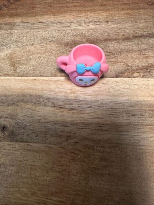 A small pink Sanrio cups resin bead with a blue bow design in the shape of a cartoon character's face sits on a wooden surface.