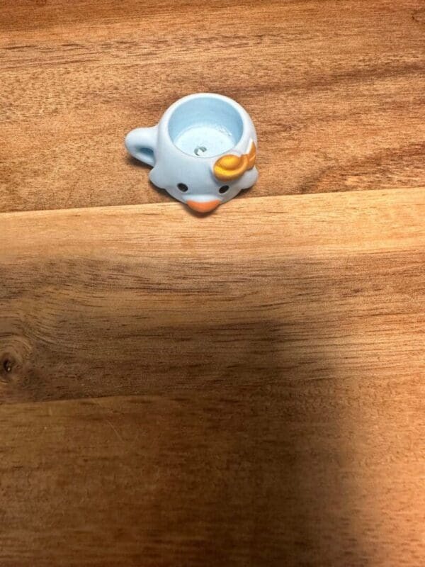 A small, blue, cartoonish Sanrio cups resin bead with a handle and an orange beak is placed upside down on a wooden surface.