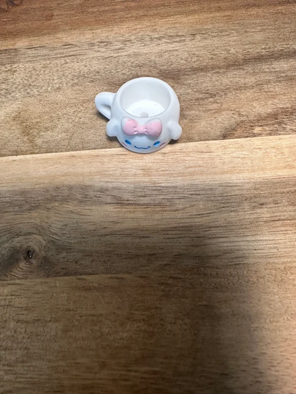 A small white Sanrio cups resin bead with handles resembling ears, featuring a smiling face with closed eyes and a pink bow, rests on a wooden surface.