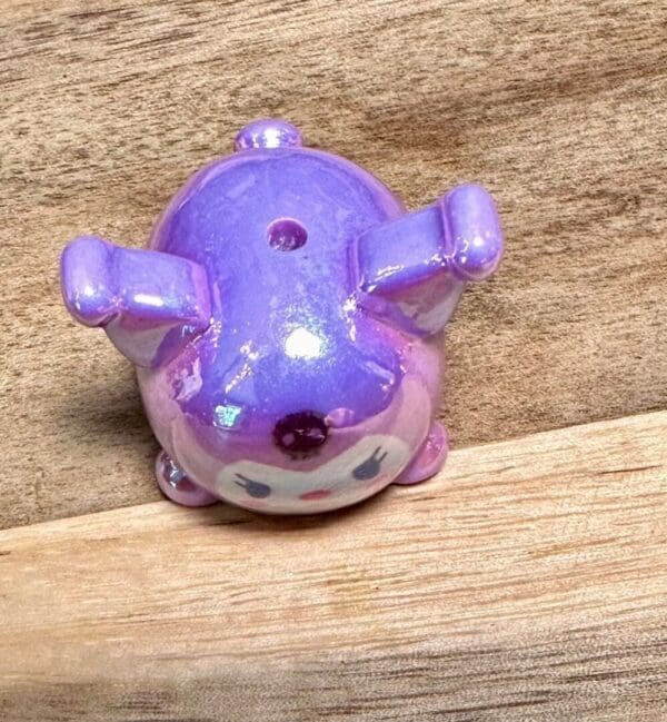 A small, glossy, purple Sanrio acrylic UV coated focal bead of a character with a round body, ears, and small limbs, lying on its back on a wooden surface.