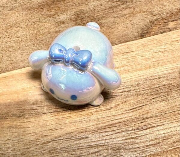 A small, glossy Sanrio acrylic UV coated focal bead of a blue character with a bow on its head, placed on a light wood surface.