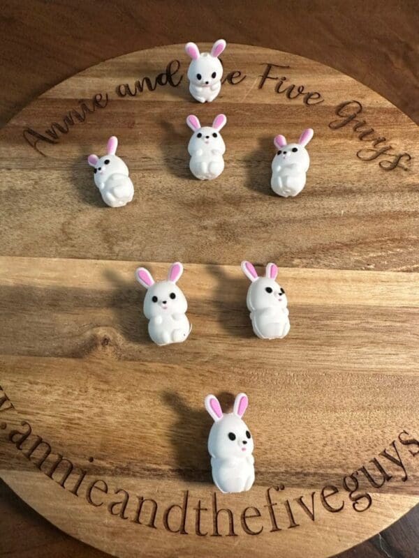 Six small, white, rabbit-shaped figurines with pink ears are arranged on a round wooden board that has "Annie and the Five Guys" engraved around the edge.