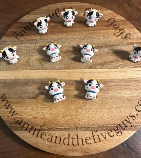 A wooden board featuring nine small, cute cow figurines arranged in a grid. The board is engraved with the text "annieandthefiveguys.com.