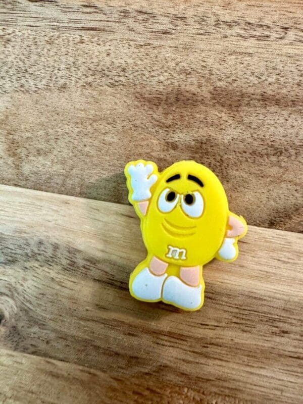 A yellow Candy silicone focal bead with a happy expression, wearing white gloves, white shoes, and standing on a wooden surface.