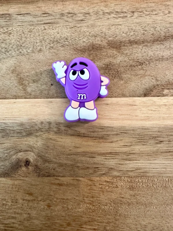 A candy silicone focal bead with white gloves and shoes stands on a wooden surface, waving with its right hand.