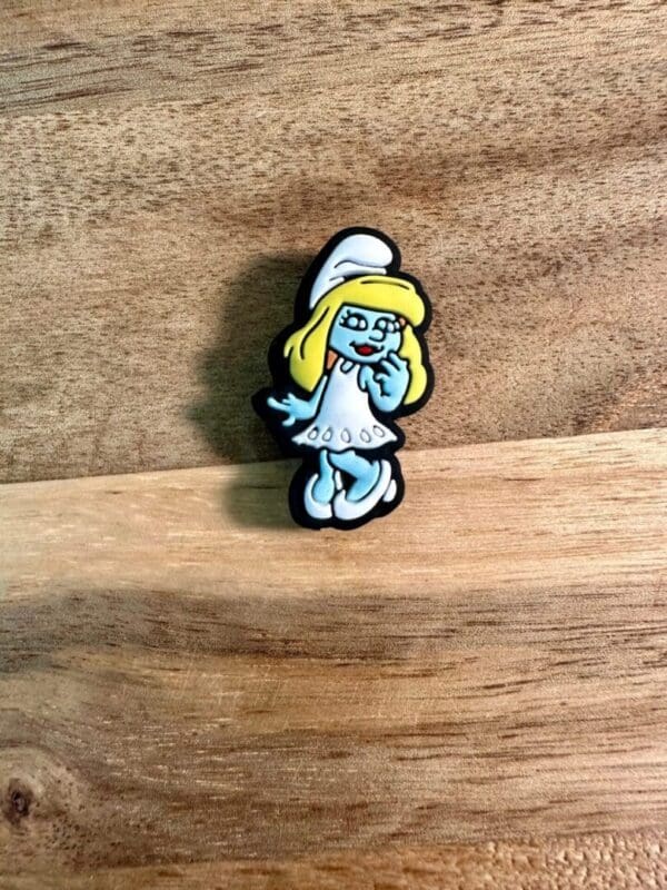 A Blue people silicone focal bead featuring a cartoon character with blue skin, yellow hair, and a white hat and dress, set against a wooden background.