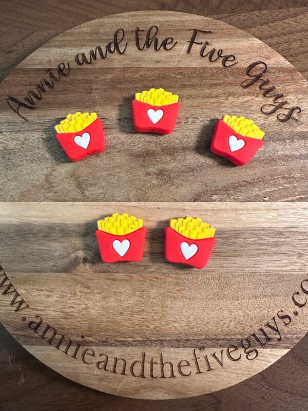 Four small fry-shaped objects with red containers and heart designs are placed on a wooden board engraved with "Sam horror guy silicone focal bead (Copy)" and a website URL.