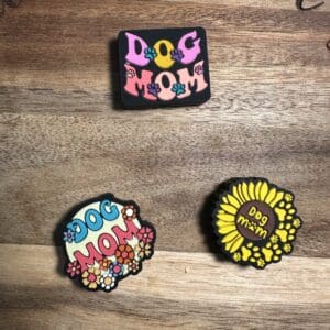 Three colorful pins on a wooden surface, each featuring the text "Dog Mom" with various floral designs.