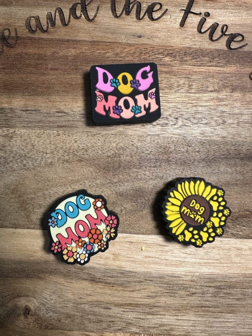 Three colorful pins on a wooden surface, each featuring the text "Dog Mom" with various floral designs.