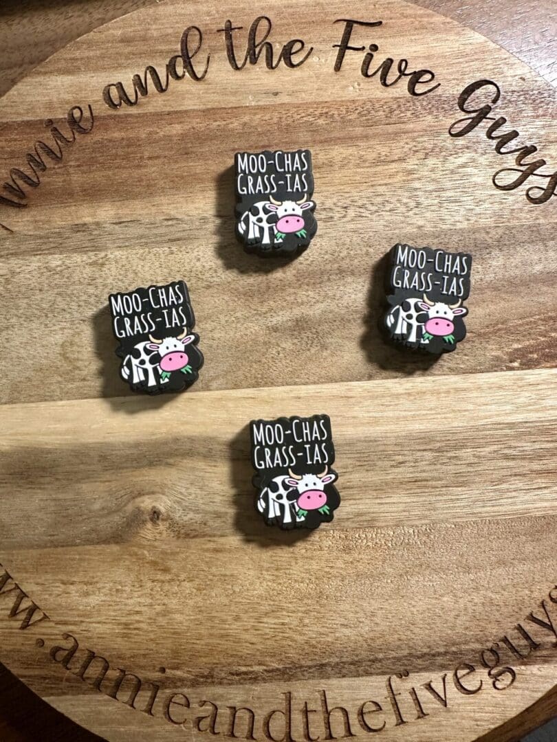 Four "Moo-Chas Gracias" enamel pins shaped like cows with the text "MOO-CHAS GRASS-IAS" are displayed on a round wooden surface with "Annie and the Five Guys" engraved around the edge.