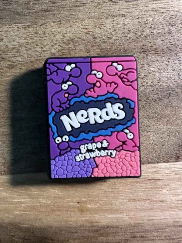 A Nerds candy container labeled "grape & strawberry," featuring a playful design of cartoon characters on a wooden surface.