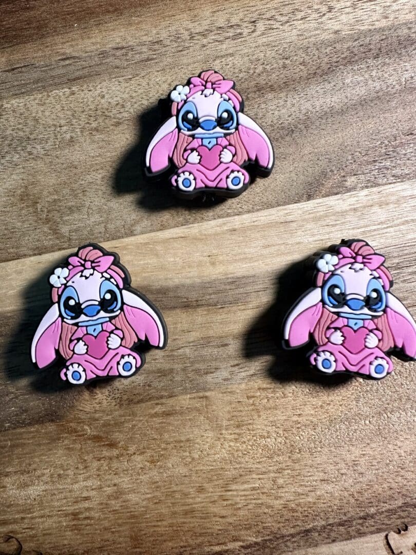 Three identical Little Pink Aliens with blue bows are arranged on a wooden surface. The aliens have large eyes and flowers on their heads.