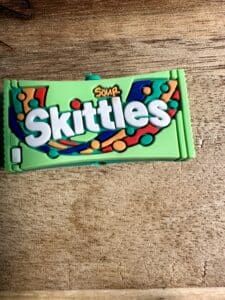 A green pack of Skittles silicone focal bead lies on a wooden surface.