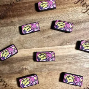 Multiple pink and yellow Hubba Bubba silicone focal beads with "Mwah Buah" printed on them are arranged on a wooden surface, which has text inscribed around the edges.