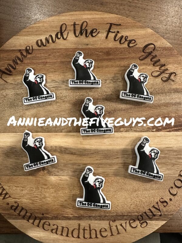 Stickers of the Exclusive Donald Trump, The OG Gangsta silicone focal bead featuring a character holding a raised fist with the text "The OG Gangsta," arranged on a round wooden board. The website "ANNIEANDTHEFIVEGUYS.COM" is displayed prominently on the stickers.