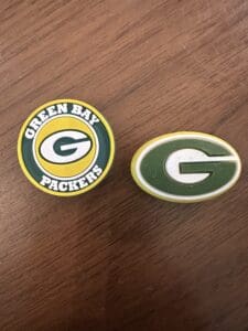 The Green Bay Silicone focal bead displays two distinct logos on a wooden surface: one showcasing a yellow 'G' with 'Green Bay Packers' text and the other featuring a simple yellow 'G' set against a green oval background.