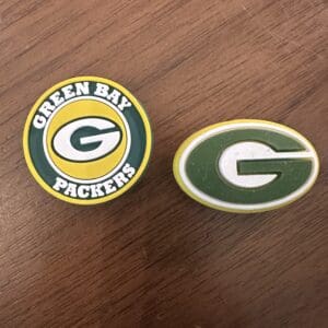 The Green Bay Silicone focal bead displays two distinct logos on a wooden surface: one showcasing a yellow 'G' with 'Green Bay Packers' text and the other featuring a simple yellow 'G' set against a green oval background.