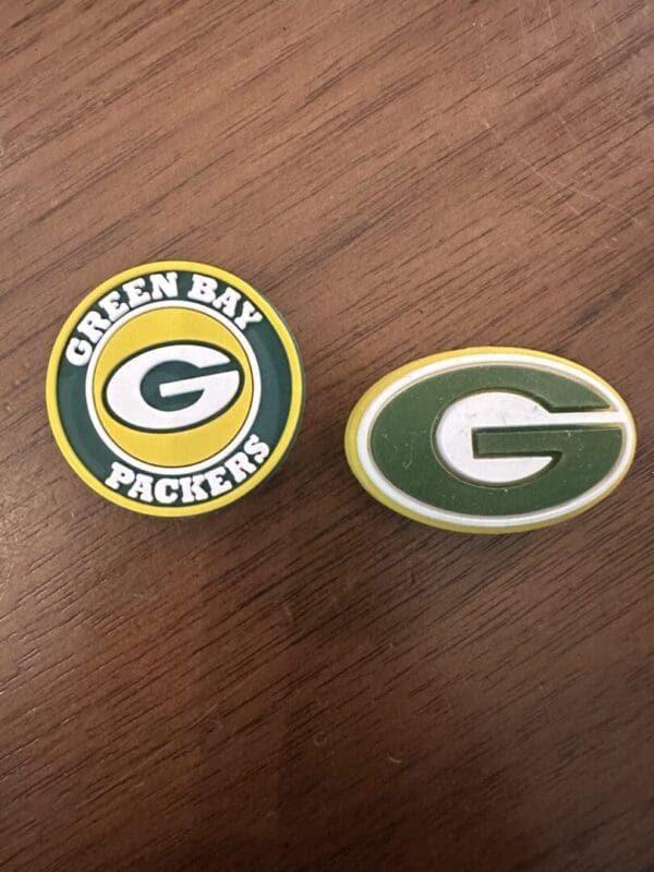 The Green Bay Silicone focal bead displays two distinct logos on a wooden surface: one showcasing a yellow 'G' with 'Green Bay Packers' text and the other featuring a simple yellow 'G' set against a green oval background.