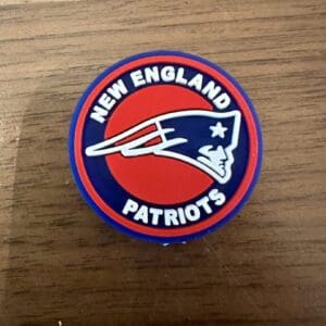 A round New England Football silicone focal bead on a wooden surface. The bead features the team's name in white uppercase letters, encircling a stylized blue and red figure of a Revolutionary War Patriot.